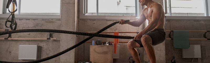 Side view of handsome physically fit male working out with heavy ropes in cross fit gym