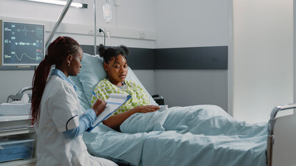 Team of african american doctors consulting patient in bed to cure illness. Medical specialists helping woman with healthcare treatment and medicine against disease in hospital ward.