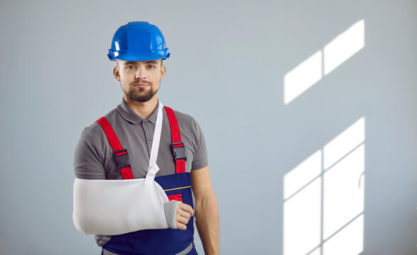 Builder gets injured at work. Portrait of man in workwear uniform, hard hat and sling immobilizer on broken arm standing on copy space wall background. Accident and injury at construction site concept