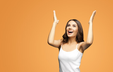 Photo of woman in white smart casual clothing raising hands up with flat palms, happy gesturing or praying, isolated on vivid brown background. Excited brunette girl looking up with smile, at studio.