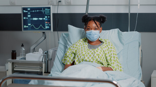 Portrait of african american patient wearing face mask, laying in hospital ward bed. Young woman with disease trying to heal during coronavirus pandemic. Sick person with protection