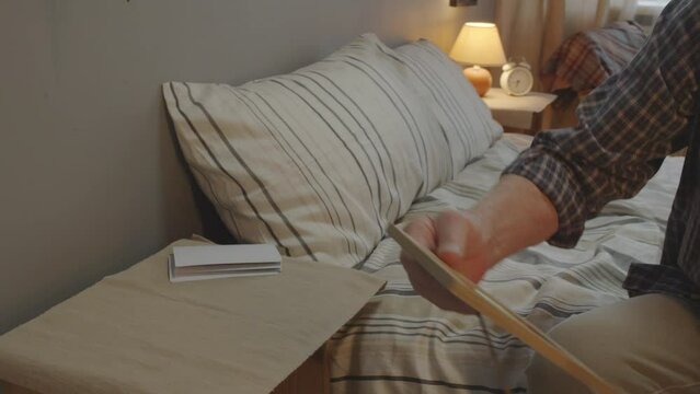 Depressed white-haired Caucasian man with mustache sitting on bed in his bedroom early in morning, looking at picture of wife in frame and thinking