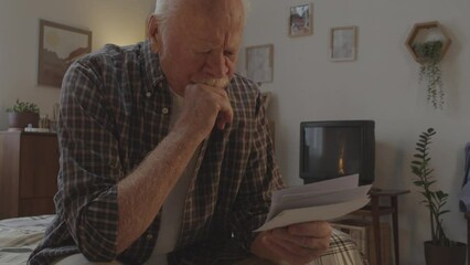 Waist-up of sad white-haired Caucasian man with mustache sitting in his bedroom early in morning, looking at old pictures and thinking