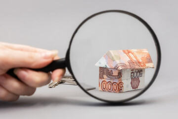 Female hand holding a magnifier glass and showing zoomed at paper origami from russian rubles. Gray background. Concept of mortgage and leasing of real estate