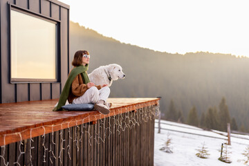 Woman sitting with dog on terrace of tiny house in the mountains. Concept of small modern cabins...