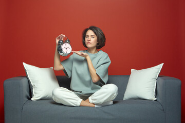 Tired young girl showing alarm clock sitting on sofa suffering sleep deprivation. Biorhythms and...
