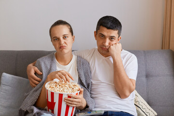 Portrait of bored sad upset wife and husband sitting on sofa with pop-corn and remote control,...