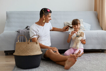 Image of Caucasian man wearing white casual style t shirt sitting on floor near sofa with child, father talking with his daughter after playing together as hairdressers.