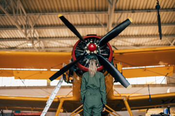 Blonde caucasian female pilot, looking at plane propeller, plane hangar.