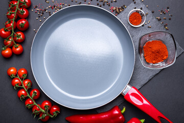Detail of an empty ceramic pan with fresh vegetables in red color around: tomatoes and peppers. Grey kitchenware with a red handle on the dark background.