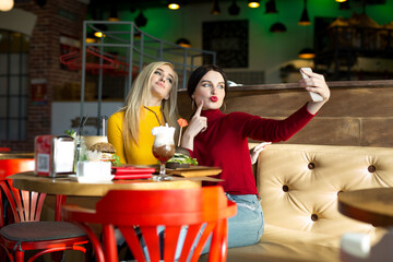 Two joyful cheerful girls taking a selfie while sitting together at cafe