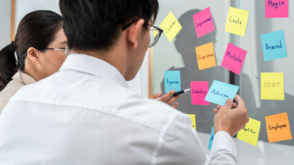 Two business people holding keyword in notes on a glass wall to brainstorm about new business project for planning marketing plan together in office