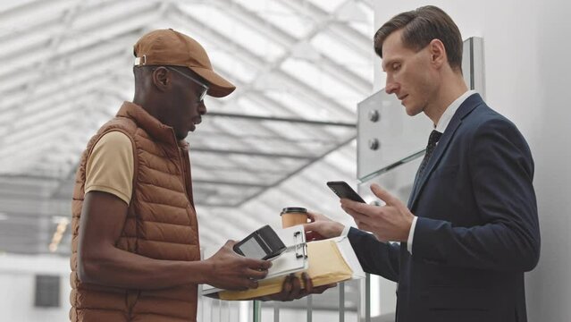 Medium shot of successful Caucasian businessman talking on mobile phone at modern business center when African American courier with terminal in hands delivering business documents in envelope to him