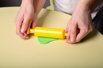 A child and his mother roll out a piece of green dough on the table with a rolling pin, close-up of hands