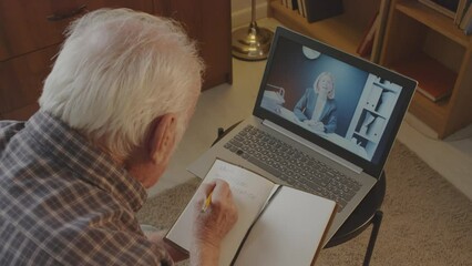 Over-shoulder of senior man sitting in his bedroom at daytime, video calling Caucasian businesswoman, pensioner taking online consultation and writing in notebook - Powered by Adobe