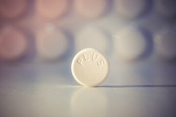 White pill with PLUS on it in selective focus. PLUS tells that the drug is more effective. There is a medicine tablet out of focus.