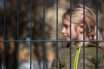 Girl child refugee behind a metal fence. Social problem of war migrants.
