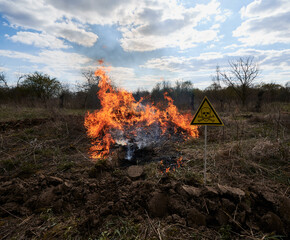 Burning dry grass and poison toxic sign under cloudy sky. Yellow triangle with skull and crossbones sign warning about danger in field with fire. Ecology, hazard and natural disaster concept.