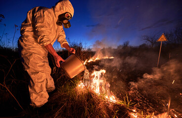 Research scientist fighting fire in field with blue night sky on background. Man in protective radiation suit and gas mask pouring water on burning grass with smoke. Natural disaster concept.