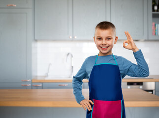 cooking, culinary and profession concept - happy smiling little boy in apron showing ok gesture over kitchen background