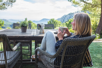 Woman drinking coffee on the terrace outdoors around mountains in sunny spring day. Girl enjoys nature and fresh air in a plaid, Health care, authenticity, sense of balance and calmness.