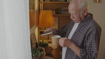 Side view of senior Caucasian man with mustache pouring alcohol from flask into mug with coffee at home in morning, drinking, looking out window