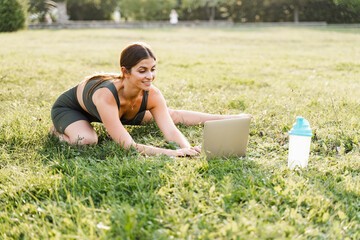 Fit girl with laptop on the green grass. Fitness trainer is chatting online with clients and...