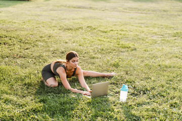 Fit girl with laptop on the green grass. Fitness trainer is chatting online with clients and...