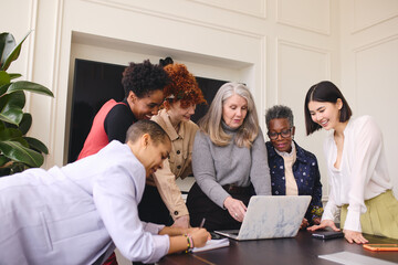 Portrait of multiethnic businesswomen looking at laptop on boardroom meeting table and discussing