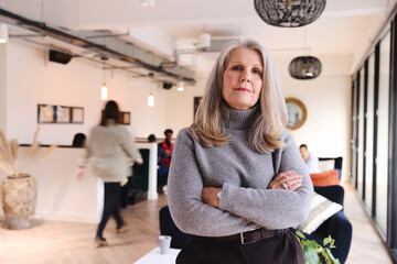 Portrait of confident senior white businesswoman with long gray hair looking at camera with arms folded in coworking space