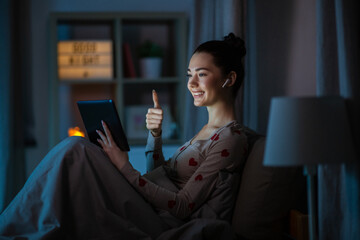 technology, bedtime and people concept - happy smiling teenage girl with tablet pc computer and earphones having video call sitting in bed at home at night and showing thumbs up