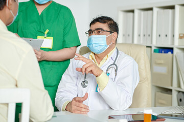 Serious general practitioner in medical mask talking to patient and explaining diagnosis