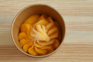 Mango sorbet in a cardboard cup stands on a wooden table. Closeup. Top-down view