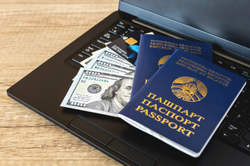 Two Belarusian passports on the keyboard of a black laptop with cash dollars and a payment card on a brown wooden table. Travel, immigration and remote work concept