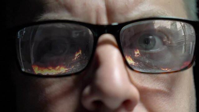 A middle-aged man watches shocking world news. Pictures of wars, fires, catastrophes are reflected in his glasses. The influence of the mass media on the human psyche in the information society.