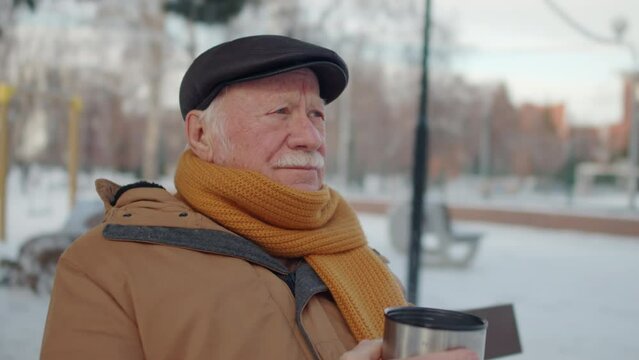 Chest up of senior Caucasian man wearing flat cap, jacket and scarf, sitting in park in winter, drinking hot tea