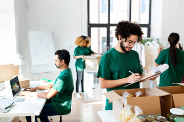 charity, donation and volunteering concept - male volunteer with clipboard packing food in boxes over international group of people at distribution or refugee assistance center