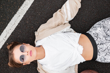 Stylish beautiful young woman with pink lips and retro sunglasses in a vintage windbreaker, white T-shirt and leggings lying and relaxing on the asphalt on a basketball court, top view