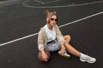 Fashionable beautiful girl with hairstyle and sunglasses in a fashionable sports jacket, leggings, white T-shirt and sneakers with a ball sitting on the street on the pavement