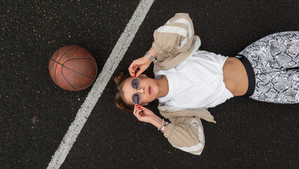 Fototapeta premium Sporty beautiful sexual woman hipster in fashionable sports clothes with a ball lying on the pavement and wearing fashion sunglasses, top view. Girl rest on the street