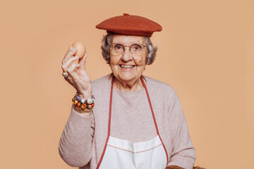 Happy smiling elderly grandmother cook holding an egg, looking at camera. Grey-haired old cook wearing apron and hat at studio over beige background. Copy space. - obrazy, fototapety, plakaty