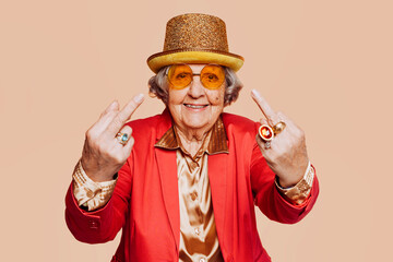 Funny stylish elderly grandmother portrait showing middle finger gesture with hand at studio. Senior old woman dressing elegant for a special event. Rebel granny over beige background. - obrazy, fototapety, plakaty