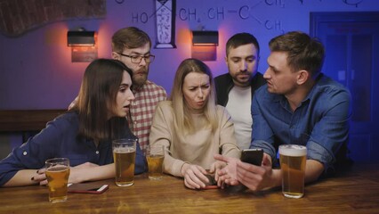 Friends in pub at bar counter with beer look at their phones. Man shows something disgusting on his phone to the friends which are grimacing Social life