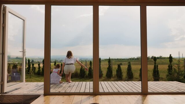 Light hair Mother with daughter barefoot goes out of house to sit at the porch. Back view