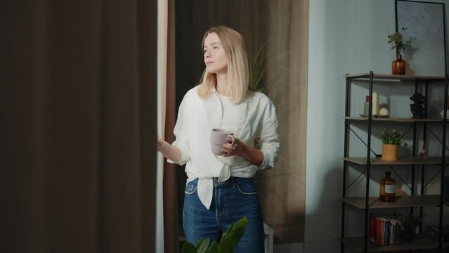 Young thoughtful beautiful woman drinking tea and looking through window while standing on windowsill at home. Footage of attractive girl tasting drink on background of modern living room. Indoors