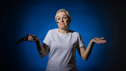 A perplexed woman with a pistol and a blue and yellow Ukrainian flag on her cheek spread her hands to the sides. Dark background.