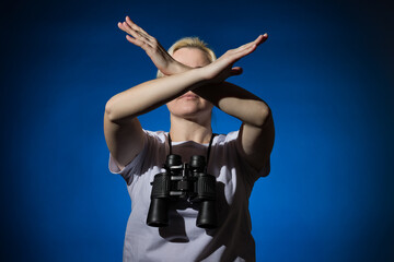 A blonde woman with binoculars around her neck shows a stop sign with her arms crossed on a dark background.
