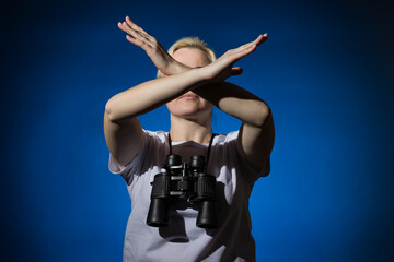 Fototapeta na wymiar A blonde woman with binoculars around her neck shows a stop sign with her arms crossed on a dark background.