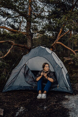 A young tourist girl is resting in a blue tent near a large tree with a mug of tea