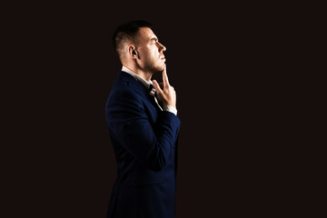 Young male businessman in a blue suit stands sideways on a dark background with copy space in hard light